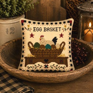 May include: A square cross-stitch sampler featuring the text EGG BASKET in black letters. The design depicts a white chicken sitting in a brown woven basket filled with colorful eggs, resting on a brown wooden table with a black heart detail.