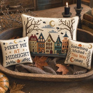 May include: Three rectangular and square cross-stitch pillows featuring embroidered designs. One pillow shows a row of colorful houses under a moon, while the others display the text "MEET ME AT MIDNIGHT" and "Something Wicked This Way Comes" with pumpkins.