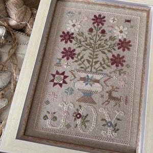 May include: A framed cross-stitch embroidery featuring a floral arrangement in a pot with the word "JOY" stitched below. The flowers are red, pink, and white, and the pot is brown with a blue and white design. The background is a beige linen fabric.