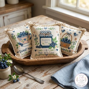 May include: Three square fabric pillows featuring cross-stitch embroidery of blueberries, a teacup, a jam jar labeled Blueberry Jam, and a basket. Each pillow has a twine bow at the top corners and the text Sweet Moments and Made With Love.