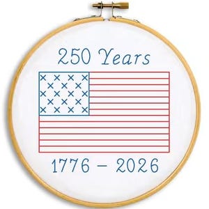 May include: A round embroidery hoop with a white fabric featuring a red and blue American flag design. The text "250 Years" and "1776 - 2026" are written in blue script. The hoop is made of wood and has a brass clasp.