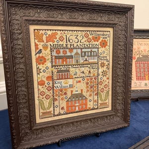 May include: A framed cross-stitch sampler featuring the text 1632, Williamsburg 1699, Middle Plantation, George Wythe, Thomas Jefferson, Patrick Henry, and King William III. The design includes houses, trees, flowers, and a bird in autumn colors.