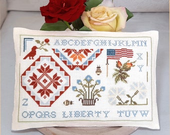 May include: A white pillow with a cross-stitch design featuring an alphabet, a bird, a flower, a bee, a flag, and a heart. The pillow is on a wooden surface with red and yellow roses.
