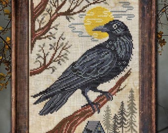 May include: Cross-stitch artwork of a black crow perched on a brown branch, set against a backdrop of a yellow moon and bare trees. A small house and evergreen trees are also visible. The artwork is framed in a dark wooden frame.