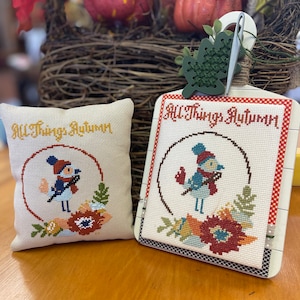 Peut inclure: Deux projets de point de croix représentant un oiseau dans une couronne avec le texte "All Things Autumn". Les projets sont sur un fond blanc avec des accents rouges, bleus et verts. Un projet est un coussin et l'autre est une pièce encadrée.