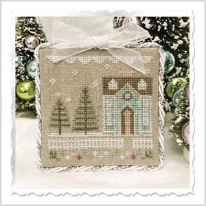 Op de afbeelding: Een vierkant, kruissteek kerstornament met een huis, twee bomen en een hek. Het ornament heeft een beige achtergrond met witte, groene en bruine steken. Bovenop zit een witte strik en de randen zijn afgewerkt met wit koord.