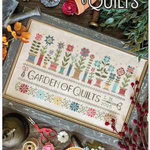 May include: A framed cross-stitch sampler titled "Garden of Quilts" with colorful floral designs and the text "Garden of Quilts". The design includes a variety of flowers in pots and spools of thread. Designed by Lori Holt.