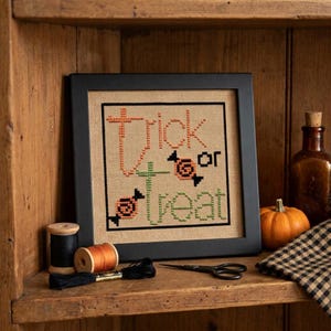 May include: A framed cross-stitch piece on tan fabric featuring the text "trick or treat" in orange and green thread, accented with two small stitched candy motifs. The artwork is enclosed in a simple black frame with a square border design.