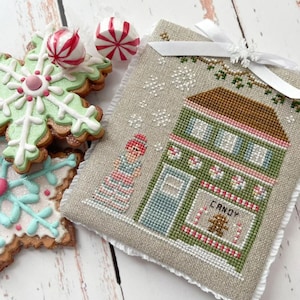 May include: A cross-stitch ornament featuring a green and pink candy shop with a gingerbread person in the window. The shop is decorated with candy canes and snowflakes. A girl in a pink and white striped dress stands in front of the shop.
