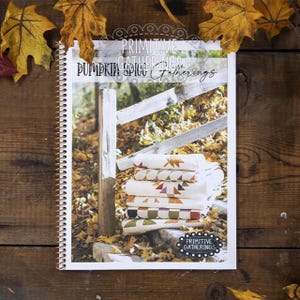 May include: A spiral-bound book titled "Primitive Gatherings Pumpkin Spice Gatherings" with a stack of patterned quilts. The quilts feature geometric and leaf designs in fall colors. Autumn leaves are scattered around the book on a wooden surface.