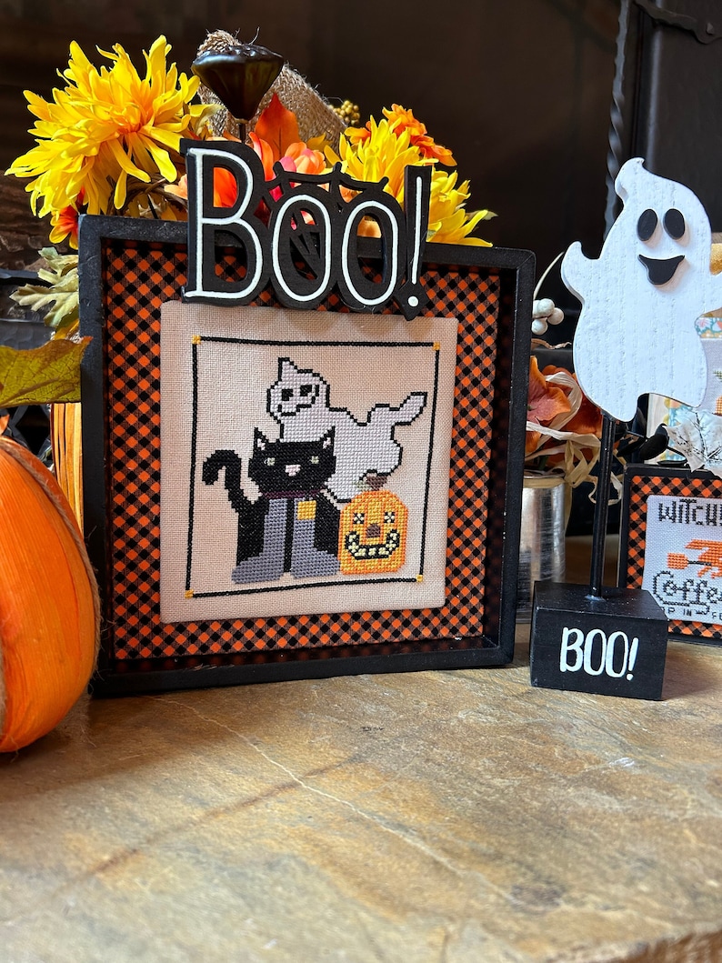 May include: A black framed cross-stitch with a black cat, a ghost, and a jack-o-lantern. The frame has a black and orange checkered border. The frame is sitting on a wooden surface with a wooden ghost and a sign that says "BOO!" in front of it.