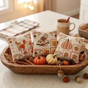 May include: Three square decorative cushions featuring cross-stitch patterns of a fox, an owl, and a pumpkin, arranged in a wooden dough bowl with miniature pumpkins, pinecones, and felt acorns. The cushions display autumn-themed motifs.