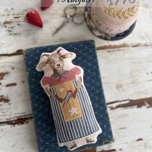 May include: A cross-stitched goat-shaped ornament from the "Animal Cracker Series + Abigail+". The goat wears a red and white patterned collar and a striped apron. The design is on a blue book with a decorative pin cushion and text that reads "1776".