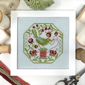 May include: A framed cross-stitch featuring a green bird perched on a spool of thread, surrounded by red flowers and green foliage. The artwork is set against a light blue background within a white frame. Wooden spools and embroidery floss are visible.