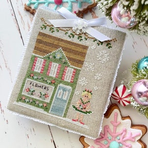 May include: A cross-stitch pattern of a small shop with a sign that reads "Flowers" and a girl in a green dress standing in front of it. The shop is decorated with a red and white striped awning and a green roof. The background is a light beige fabric with a white lace border.