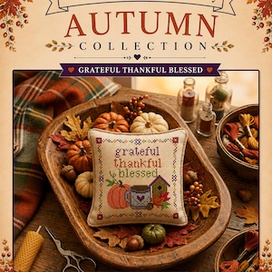 May include: A square cross-stitch sampler featuring the text grateful thankful blessed, a pumpkin, a mug, and a birdhouse. The sampler is displayed in a wooden bowl with miniature pumpkins, acorns, and autumn leaves, surrounded by embroidery floss.