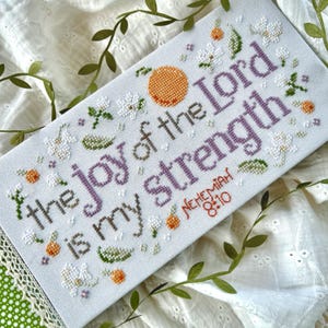 May include: A cross-stitch piece with the text "the joy of the Lord is my strength" in purple. The design includes orange fruit, white flowers, and green leaves. The scripture reference "Nehemiah 8:10" is in red.
