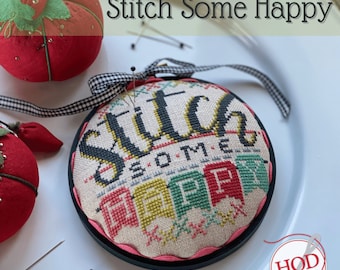 May include: A cross-stitch embroidery hoop with a black frame and a white fabric background. The embroidery features the text "Stitch Some Happy" in black and yellow lettering with colorful flags below. The hoop is sitting on a white plate with two red pincushions.