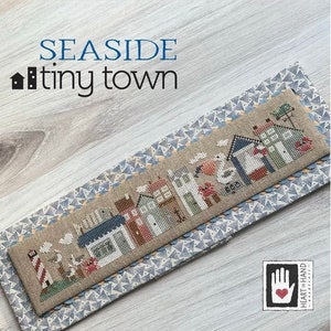 May include: A cross-stitch pattern of a seaside town with houses, a lighthouse, and a seagull. The pattern is on a beige fabric with a blue and white border. The text "SEASIDE tiny town" is above the pattern. The Heart in Hand logo is in the bottom right corner.