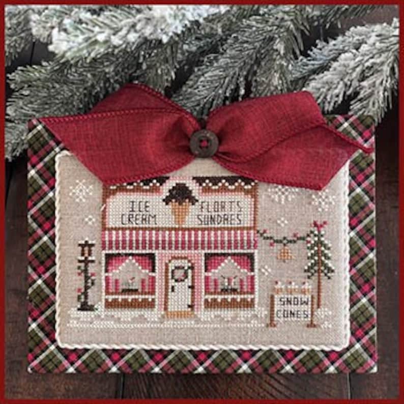 Op de afbeelding: Kruissteekpatroon van een kleine, rood-witte winkel met een bord waarop "Ice Cream & Florets Sundaes" staat. De winkel heeft een rode deur en twee ramen met een kleine kerstboom en een bord waarop "Snow Cones" staat. Het patroon is omlijst met een rood-groene ruit.