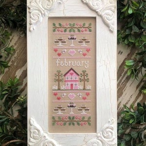 May include: White framed cross-stitch embroidery with a pink house, trees, hearts, and the word "February" in a brown font. The embroidery also features two cakes with pink frosting and brown chocolate.