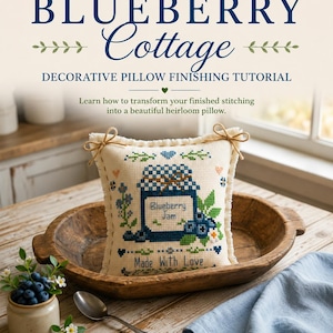 Puede incluir: Un cojín decorativo cuadrado con un diseño de punto de cruz de un tarro etiquetado como Blueberry Jam y el texto Made With Love. El cojín tiene un borde deshilachado y está atado en las esquinas superiores con un cordel marrón fino.