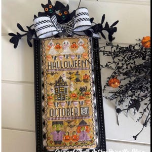 May include: A rectangular cross-stitch wall hanging featuring Halloween motifs including ghosts, a haunted house, a black cat, Frankenstein, and a witch. The text reads HALLOWEEN and OCTOBER 31. It is framed in black with a black and white bow.