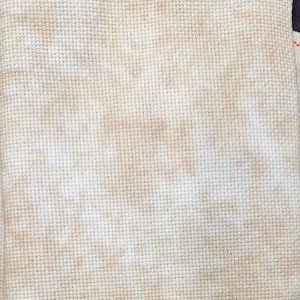 May include: Close-up of a piece of off-white cross-stitch fabric with brown mottled markings. The fabric has a grid-like pattern, and the brown markings are irregularly distributed across the surface.