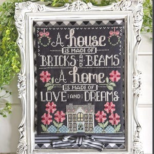 May include: A framed cross-stitch sampler on dark gray fabric featuring the text A house is made of bricks and beams a home is made of love and dreams, decorated with pink flowers, green leaves, and a small house illustration with a striped bow.