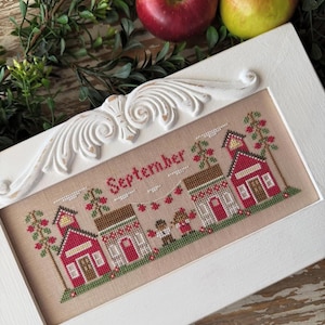 May include: A framed cross-stitch piece with the word "September" stitched in red. The design features red buildings, trees with red berries, and small figures. The frame is white with ornate detailing, and two apples are visible.