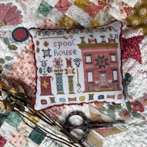 May include: A square pincushion featuring a cross-stitch design of a red house, various spools of thread, and the text "spool house" in dark lettering. The pincushion has a ruffled red border and is displayed on a patchwork quilt background.