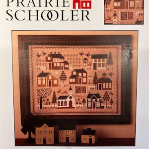 May include: A cross-stitch pattern for a village sampler, featuring a church, houses, and trees. The pattern is framed in a brown frame and is titled "Village Sampler". The pattern is from "The Prairie Schooler" book number 15.