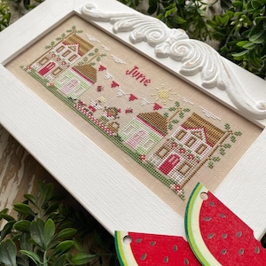 May include: A white framed cross-stitch with a scene of houses and a red heart garland. The cross-stitch is titled "June". Two watermelon slices are in the foreground.