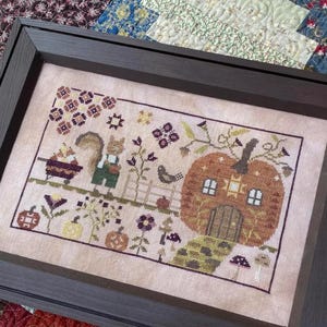 May include: Cross-stitch artwork in a dark wood frame. The design features a squirrel in overalls, a pumpkin house, and various autumn elements like pumpkins, flowers, and mushrooms. The color palette includes shades of brown, green, and orange.
