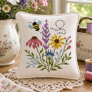 May include: A square white fabric pillow featuring a cross-stitch design of a bumblebee flying above a cluster of colorful wildflowers including purple lavender, a yellow sunflower, a pink coneflower, and various green foliage and blue stems.