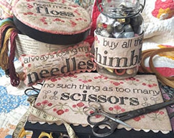 May include: A collection of needlework supplies, including a jar of buttons, a needle minder, a floss container, and a pair of scissors. The image includes the text "Heartstring Samplery", "Need more floss", "Always losing my needles", "Buy all the thimbles", "No such thing as too many scissors", and "For the love of stitching".