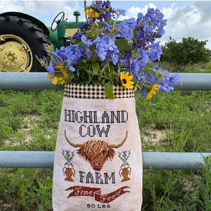 May include: A beige fabric bag with the words "HIGHLAND COW FARM Fine Feed" and a Highland cow illustration. The bag is filled with blue and yellow flowers and is hanging on a metal fence. The bag is labeled "50 LBS".