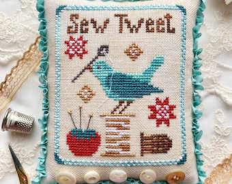 May include: A cross-stitch pillow with a blue bird holding a needle and thread, surrounded by sewing supplies. The pillow is titled "Sew Tweet" and has a white background with colorful accents.