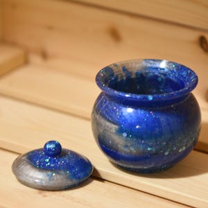 May include: A blue and silver glitter resin jar with a lid. The jar is round and has a smooth, glossy finish. The lid is also round and has a small knob on top.