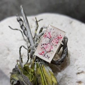 Könnte beinhalten: Ein silberner Ring mit rechteckiger Oberfläche, der ein Blumendesign aus rosa Rosen zeigt. Der Ring ist vor einem Hintergrund aus grauen, grünen und silbernen dekorativen Elementen platziert, was auf ein botanisches oder naturnahes Thema hindeutet.