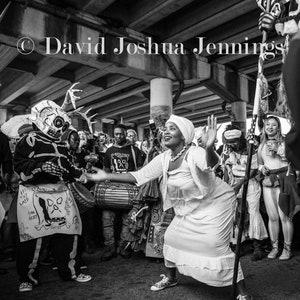 May include: A black and white photograph of a group of people in costume, some wearing masks and others in white clothing. The people are dancing and playing drums under a bridge. The photo is taken from a low angle, looking up at the people.