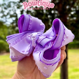 Puede incluir: Un lazo grande para el cabello de color lavanda, con un diseño en capas y detalles de perlas. El lazo presenta un borde con volantes y se sostiene en una mano. El texto "Bowtique on Main Street" es visible.