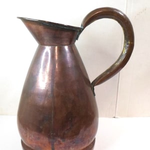 May include: A copper jug with a wide mouth and a curved handle. The jug has a tarnished finish and a rounded body.