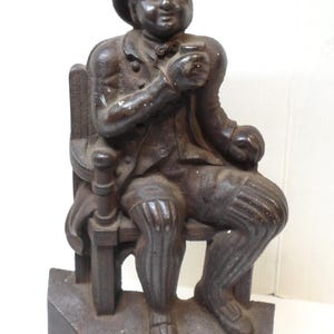 May include: A dark brown cast iron statue of a man seated in a chair, wearing a hat and holding a cup. The figure is dressed in a jacket, breeches, and shoes. The statue sits on a rectangular base. The statue is a decorative item.