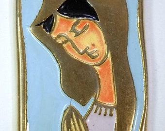 May include: A rectangular metal pendant featuring a cloisonné enamel design of a figure with long hair and hands pressed together in prayer. The figure is rendered in shades of orange, white, and black against a light blue background with gold trim.