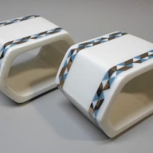 May include: Two white ceramic napkin rings with a geometric pattern in blue, brown, and silver. The rings are shaped like a hexagon.