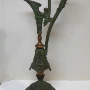 May include: A tall, slender metal ewer featuring a weathered green patina finish, a long curved handle, and a flared spout. The body displays intricate relief detailing, a central copper-toned band, and a circular dark brown base.