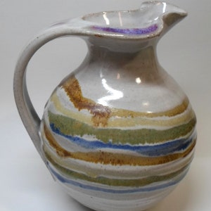 May include: A ceramic pitcher with a curved handle and a spout. The pitcher is white with horizontal stripes in shades of brown, green, blue, and yellow. The rim of the pitcher has a purple accent.
