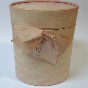 May include: A cylindrical, light pink gift box with a matching lid. The box is adorned with a delicate floral pattern and tied with a soft pink ribbon. A small tag is attached to the ribbon. The box is designed for gifting.