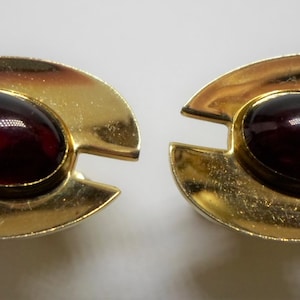 May include: A pair of gold-toned clip-on earrings. Each earring features a dark red, oval-shaped stone set in a gold-colored, crescent-shaped frame. The earrings have a vintage aesthetic and a polished finish.